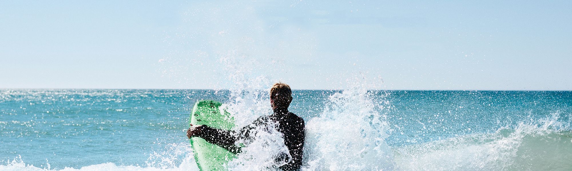 Ein Mann rennt mit seinem Surfbrett durch eine Welle ins Meer bei klaren Himmel und Sonnenschein