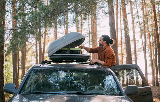 Vermietservice für die Urlaubsreise beim Autohaus-SCHADE. Ein Mann klappt seine Dachbox auf dem Dach des Fahrzeuges auf. Das Fahrzeug ist dunkelgrau. Er steht bei Sonnenschein im Wald.
