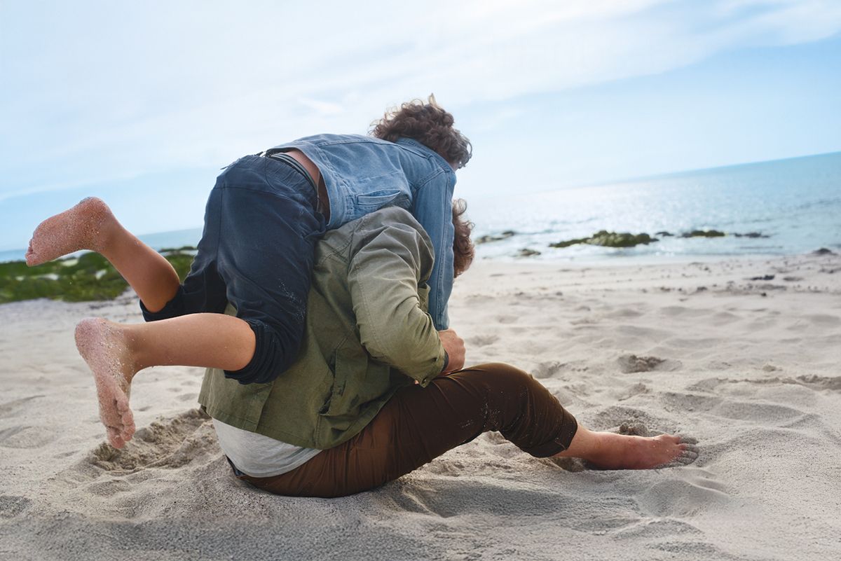 Ein Elternteil spielt mit einem Kind am Strand. Beide sitzen im Sand, das Kind klettert lachend auf den Rücken des Erwachsenen. Im Hintergrund das ruhige Meer und ein heller Himmel – ein Moment voller Sommerfreude und Familienzeit.