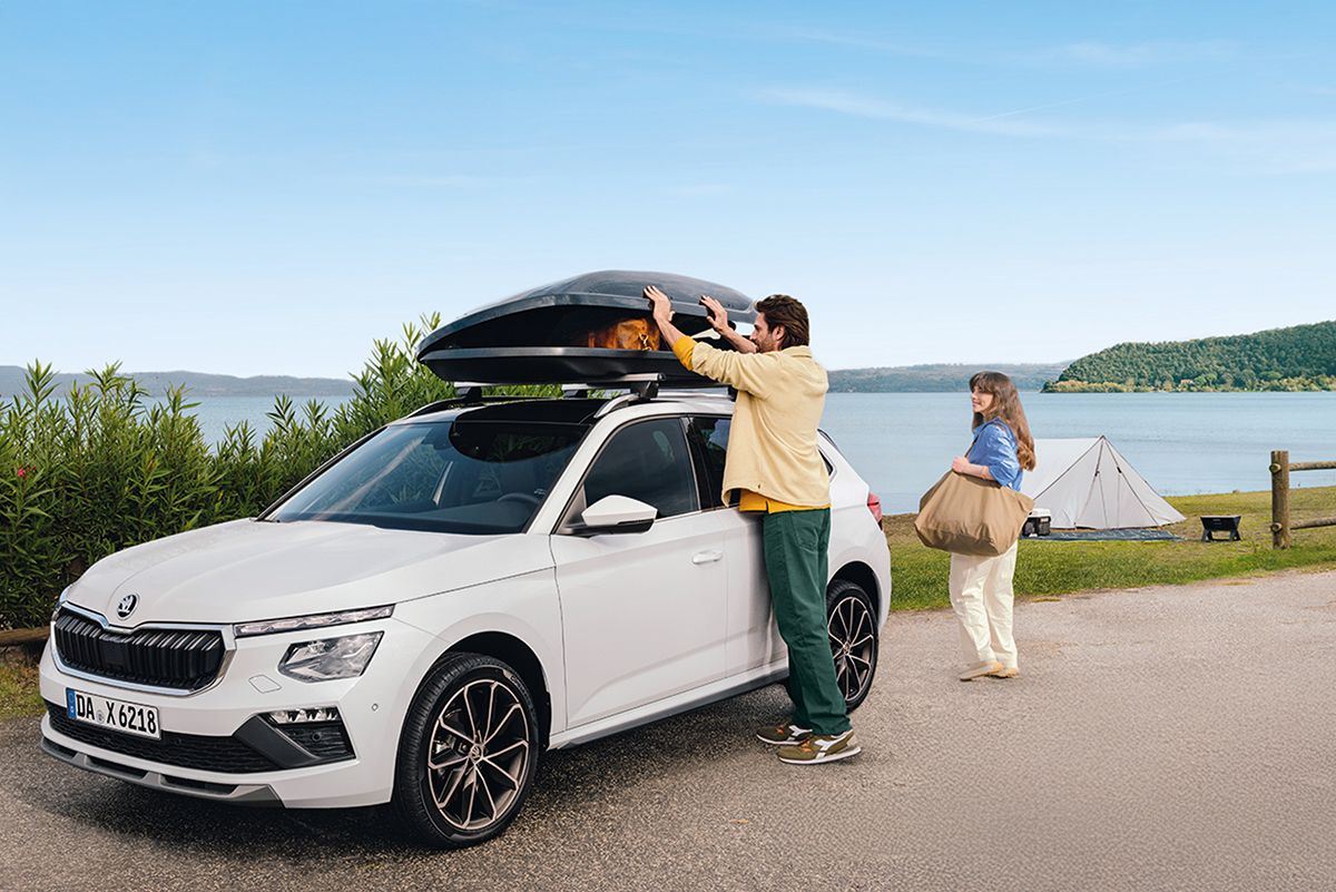 Ein Mann verstaut Gepäck in die Dachbox eines weißen Škoda vor einem See. Eine Frau mit Tasche steht daneben – im Hintergrund Zelt und Natur. Urlaubsstimmung beim Camping.